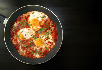 Shakshuka - poached eggs in tomato sauce, onion, pepper and spices in iron pan on dark wooden table with copy space. Famous traditional Arabic and Israeli breakfast - chakchouka. Top view