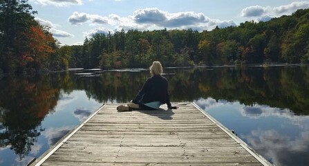 Female Sitting on the Edge of a Wooden Dock with Stunning Views of Autumn Trees and Lake Reflections