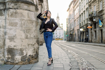 Full length portrait of young brunette Asian woman in jeans and stylish black leather jacket, sexy posing near old stone wall of the building, outdoor summer shot