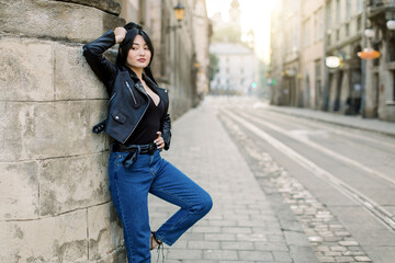 Young elegant mixed raced brunette woman model in jeans and fashionable leather jacket, standing in the city leaning on vintage wall. Attractive fashion model girl in the street. City style