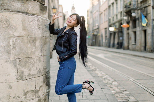 Beautiful young Asian model woman with ponytail hair, in fashionable casual black leather jacket and denim, and heels, standing near old building in ancient city center and showing peace victory sign