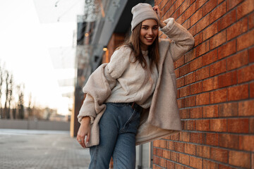 Happy young woman model in a vintage hat in a stylish knitted sweater in a faux fur coat is stands and smiling near a brick building in the city. Joyful girl in trendy clothes is resting on the street