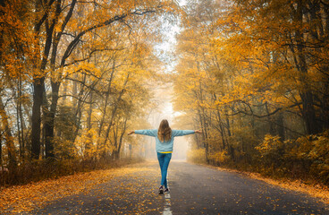 Happy young woman on the road in beautiful autumn forest in fog at sunset. Landscape with alone girl, road, orange trees. Empty roadway through foggy woodland in fall. Travel and adventure in Europe
