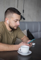 Young man in casual clothes listening to music in wireless earphones while using smartphone in cafe.
