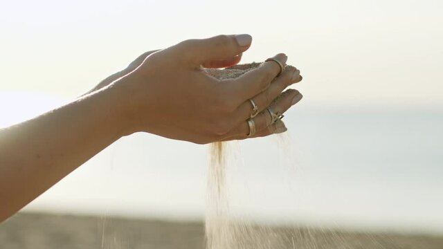 Close up of woman pouring sand running through fingers with boho rings slow motion at beach with sun flare. Beautiful scene of young lady playing with sand on sunrise or sunset.