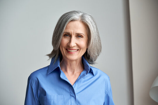 Smiling sophisticated mature grey-haired woman standing on grey wall background at home. Happy elegant middle aged old lady professional businesswoman entrepreneur posing in office, headshot portrait.