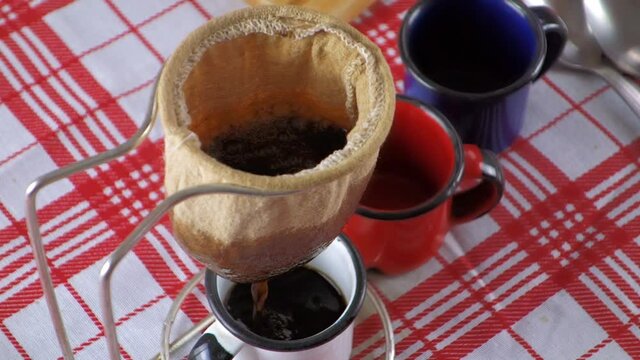 Coffee being filtered through a cloth strainer in enameled cups and homemade bread in the background on a checkered tablecloth. Making coffee with an cloth strainer also known as Mariquinha in Brazil