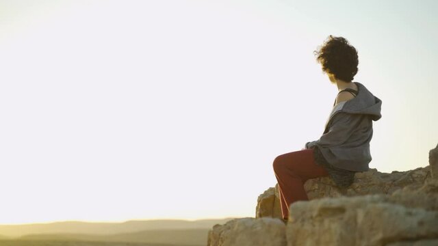 Desert sunset or sunrise with young woman sitting and watching the horizon. Hippie dressed girl or backpacking traveller sitting on a rock on a cliff mountain top. Pensive and contemplating in nature.