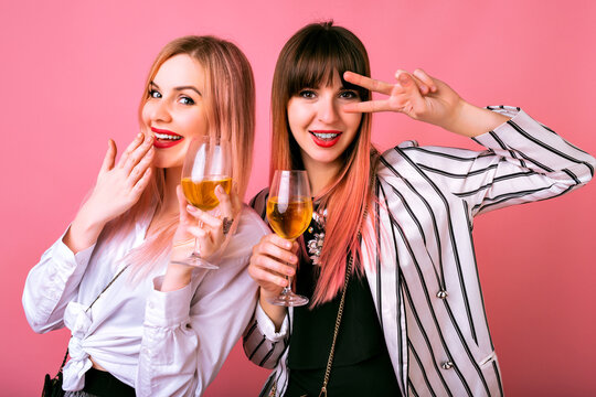 Positive indoor studio portrait of two stylish elegant pretty women having fun on party, drinking tasty champagne and dancing, cocktail evening outfits and pink background.