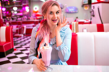 Indoor image of pretty young elegant woman enjoying her tasty sweet strawberry milk shake at retro vintage American restaurant, neon design, cute pastel dress, pink hairs and accessories.