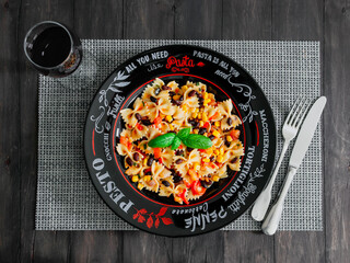 Farfalle pasta with vegetables.

Farfalle pasta with vegetables in a plate with red wine and cutlery on a black wooden table, top view close-up.