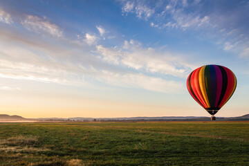 Hot Air Balloon in colorful rainbow stripes begins ascent over farm field as sun rises blue cloudy sky