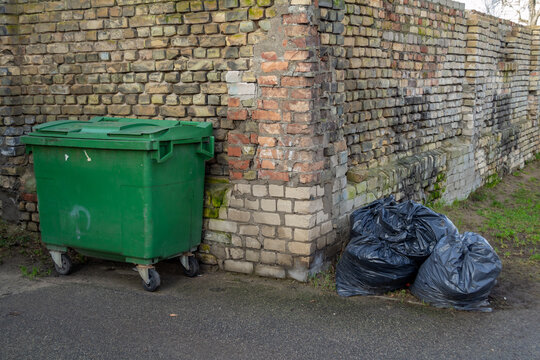 Green container and pile of garbage bags. Lots black garbage bag with street and old brick wall background. Riga, Latvia