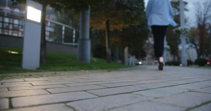 Woman in blue shirt kicks e-scooter to ride and disappears in evening city park slow motion close backside low angle shot