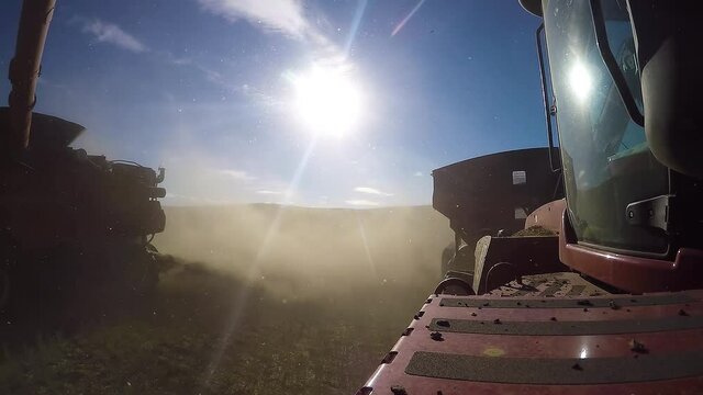 POV grain cart tractor: farm combine harvester unloading grain to tandem driving machine in rural farm countryside land on bright sunny blue day with sun rays beaming in sky, close up