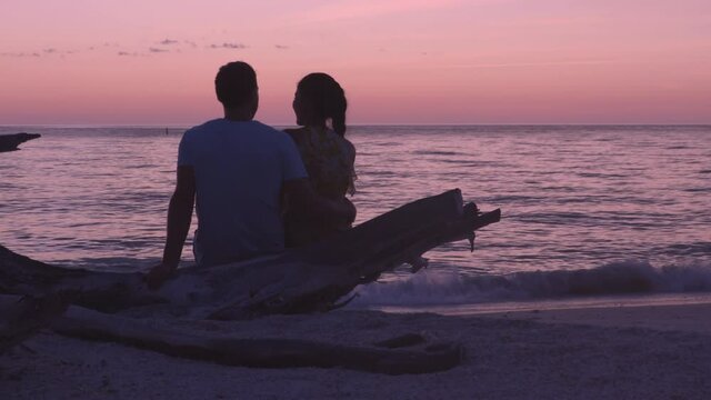 Romantic beach sunset couple honeymoon - Lovers enjoying watching sunset on summer travel destination sitting on tree trunk by the ocean. From Lovers key, Florida near Fort Myers Beach, USA.
