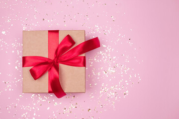 Gift box with red bow on festive pink background with golden sparkles around and copyspace for your text. Flat lay style. Christmas, New Year, Valentines Day or birthday celebration concept