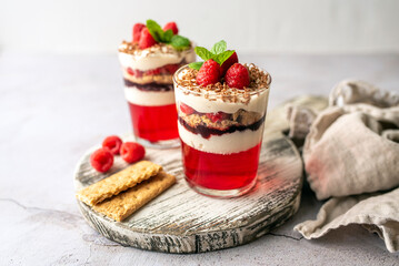 Dessert in a glass cup, with Jello covered in yogurt and topped with fresh raspberries. Mini dessert on light concrete background. Fruit parfait with jello and jelly.  - obrazy, fototapety, plakaty