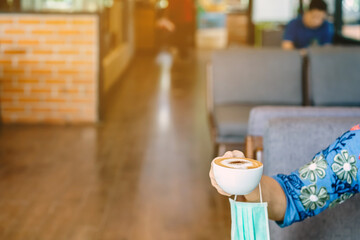 Hot Cappuccino coffee with protective mask to prevent Corona Virus (Covid-19) hanging on woman arm while take off mask to drink coffee at coffee shop. New normal lifestyle concept. Selective focus