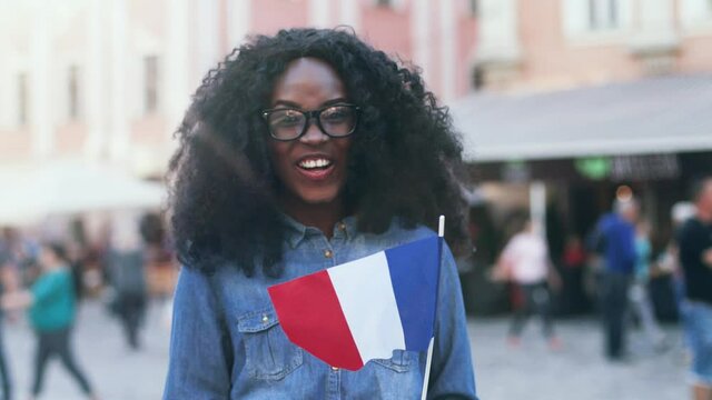 Portrait of young happy exchange student with lush curly hair. Beautiful girl in denim shirt is smiling happily and waving the flag of France on outside.