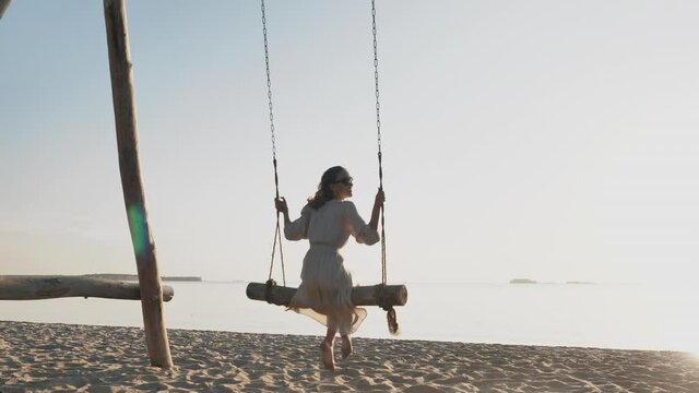 Young happy joyful girl flies swaying on wooden swings near sea on paradise beach in sunshine alone. Enjoyment of freshness, serene relax, freedom, summer vacation on dream resort by sunny day outdoor