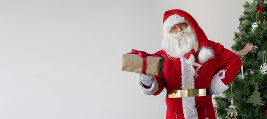 santa claus is standing near the christmas tree, holding a box with a gift.
