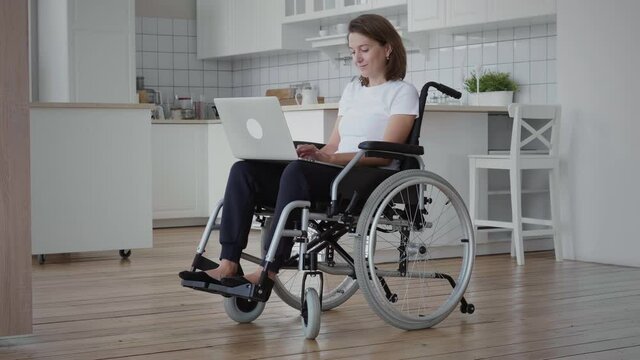 Mid shot of young brunette disabled woman sitting on wheelchair and using laptop in the living room. Freelancer, young writer or student work with laptop