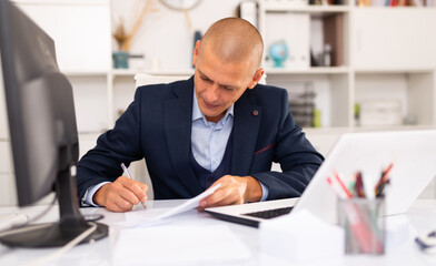 Fototapeta premium Focused thoughtful young man working with laptop and documents in office
