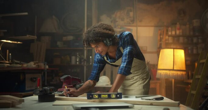 Creative curly Latin carpentry master working at cozy workshop drawing furniture project, confident male carpenter in apron and anti-noise headphones using pencil to mark measurements on work-table