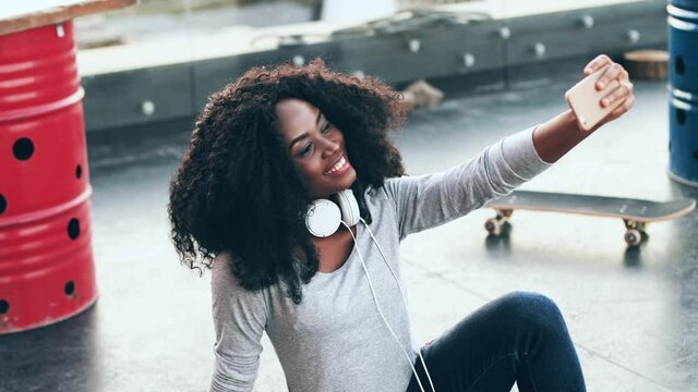 Cheerful young woman with curly hair sitting on terrace and taking selfie on her smartphone. Beautiful african american woman sitting on roof posing at mobile camera holding in outstretched hand.