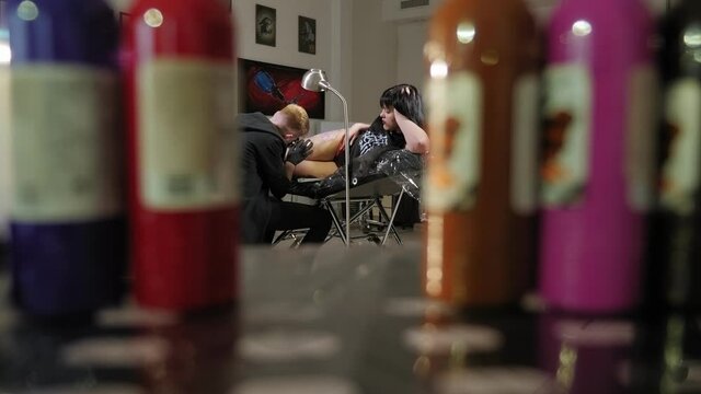 A man tattoo artist is tattooing on the leg of a young woman in a modern tattoo parlor, in the foreground are multi-colored inks.