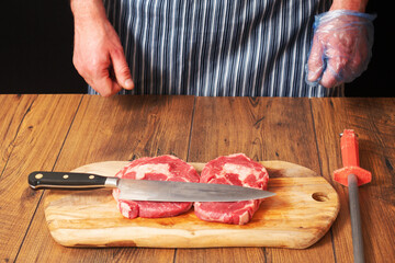 Knife over two fresh rib eye steaks on a wooden board. Professional butcher in black and white apron.