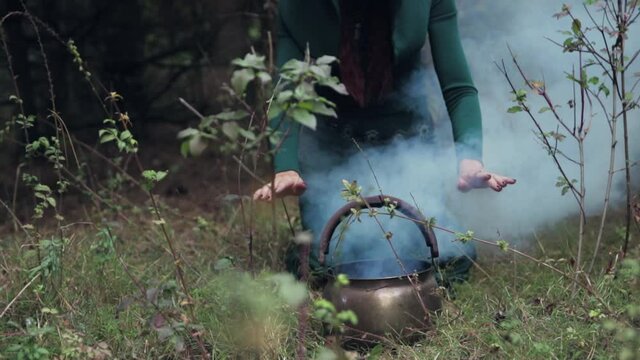 A witch in a green hooded dress conjures her hands over a cauldron of potion in the forest. Blue smoke comes out of the boiler. Black magic