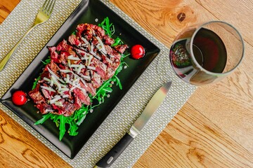 Tagliata di carne con rucola, parmigiano, aceto balsamico e pomodori fotografata dall'alto in un piatto nero e con bicchiere di vino rosso