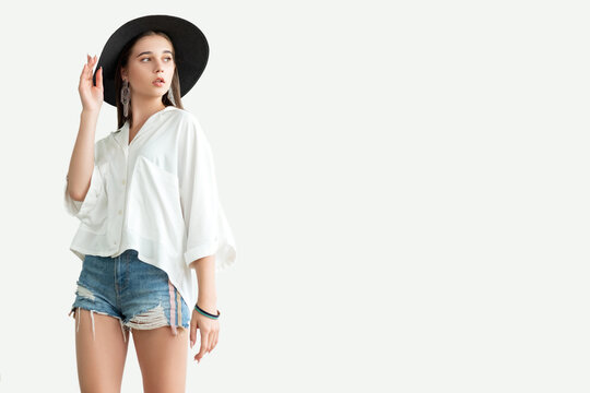 Summer outfit. Trend stylish look. Young woman in white shirt jeans shorts and wide-brimmed black hat looking copy space isolated on neutral. Casual eleganc. Advertising background