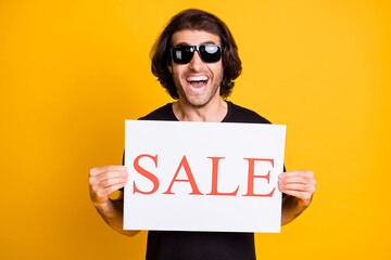 Photo of young man hold demonstrate paper poster open mouth wear t-shirt sunglass isolated yellow...