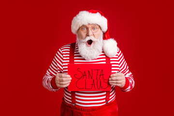 Portrait of his he nice handsome amazed stunned experienced bearded St Nicholas holding in hands card saying Santa Claus event organization isolated bright vivid shine vibrant red color background