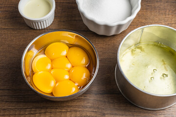 Raw ingredients in containers for baking, making a cake, or other desserts. Raw separated egg yolks, egg white, white sugar, cream. On a wood table top