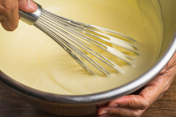Whisking mixing smooth light yellow cake mixture with a whisk in a metal bowl on top of wood table...