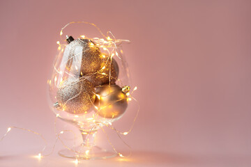 In a glass vase on a pink background are Golden Christmas balls in sequins and a garland with yellow light. Christmas home decoration. space for text