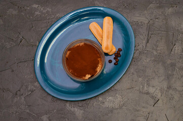 Dessert of italian cuisine tiramisu with cocoa and savoyardi cookies in a glass bowl on blue plate, gray concrete background, top view