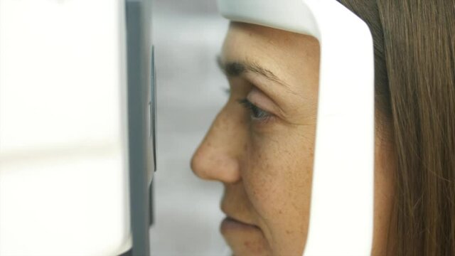Close-up portrait of woman during test of refractometer machine, side view. Eye exam for young woman at optical store.