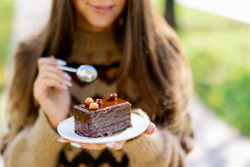 Smiling caucasian female model eating fancy slice cake in outdoor on background of green park,...