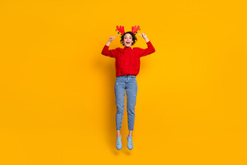 Full length photo of pretty funky lady jump high up chilling christmas party showing her head decor wear headband x-mas horns red knitted sweater jeans shoes isolated yellow color background