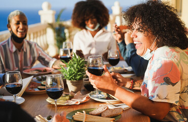 Young multiracial people eating and drinking red wine while wearing protective masks - Happy...