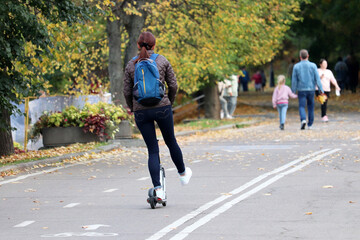 Woman rides an electric scooter on a city street, rear view. Riding e-scooter in autumn