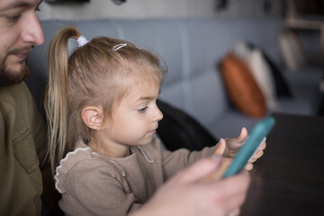 young father and his daughter watching funny video in smartphone  in cafe. man and little girl are sitting at a table in a cafe and everyone is looking into his phone. Lack of f live communication.