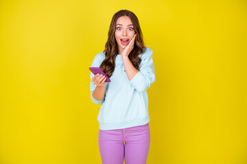 Photo of cute excited girl hand face hold mobile recieve text friend got university degree invite bachelour party wear blue sweater violet trousers isolated vivid yellow color background