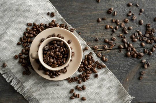 Coffee cup filled of fresh arabica or robusta coffee beans with scattered coffee beans on a linen textile and wood table.