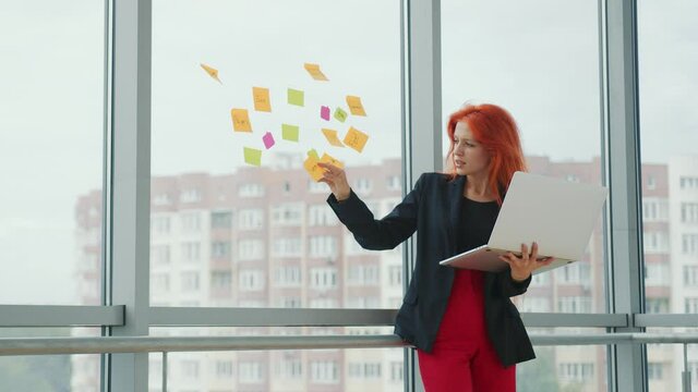 Office work with laptop and stickers. Young red-haired woman holds a laptop in her hand and takes a sticker from the window and examines it. Business lady in the office at work.